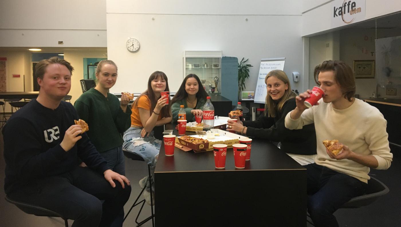 Seks ungdommer som sitter rundt et bord og spiser pizza og drikker brus
